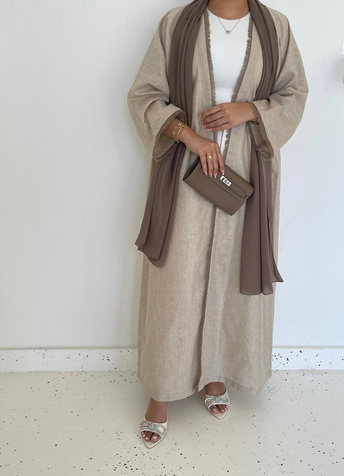 Person wearing a beige long coat with a brown scarf and holding a matching clutch against a white background
