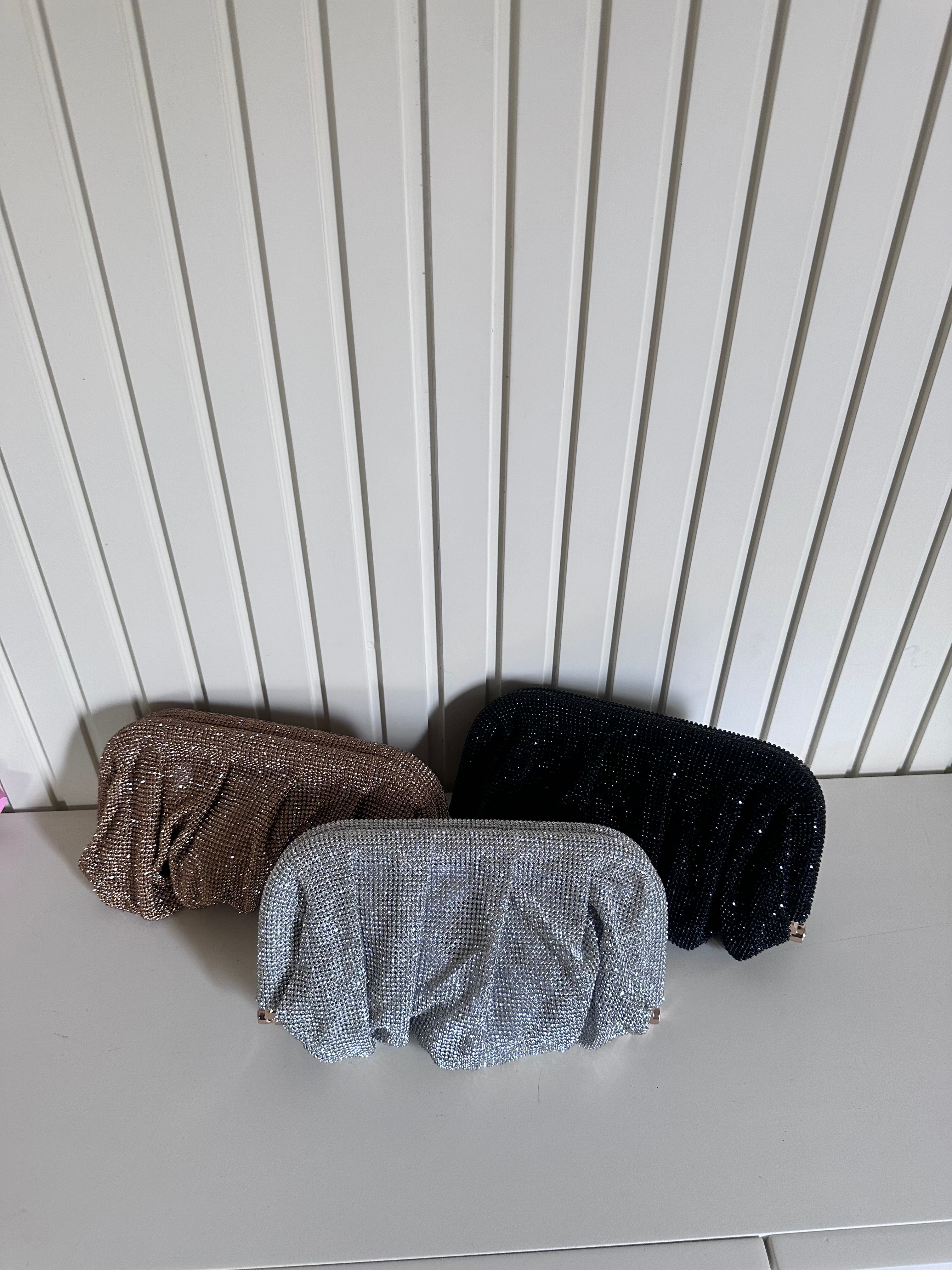 Three glittery pouches in brown, gray, and black on a light surface with a white paneled wall background.
