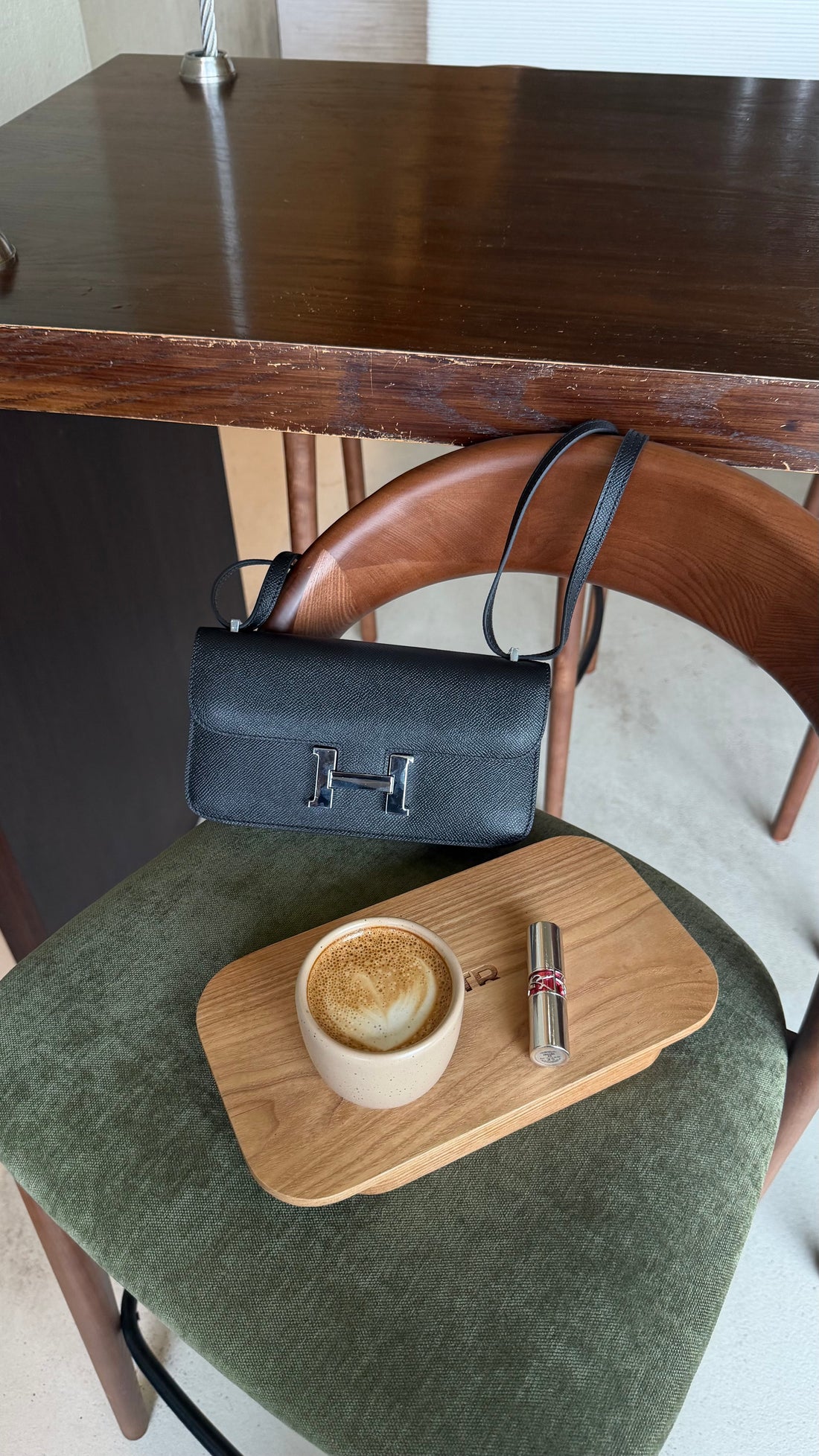 Black handbag on a chair with a wooden tray holding a cup of coffee and a lipstick.