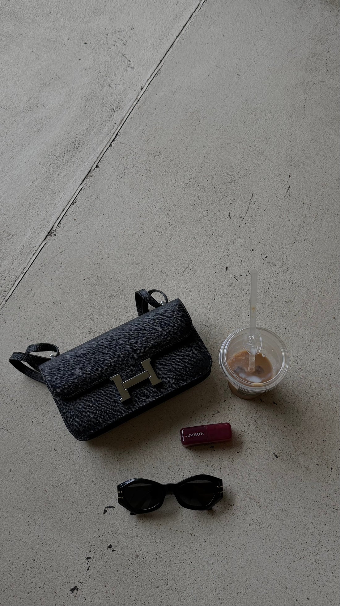 Black handbag with gold 'H' logo, sunglasses, and a small container on a concrete floor.