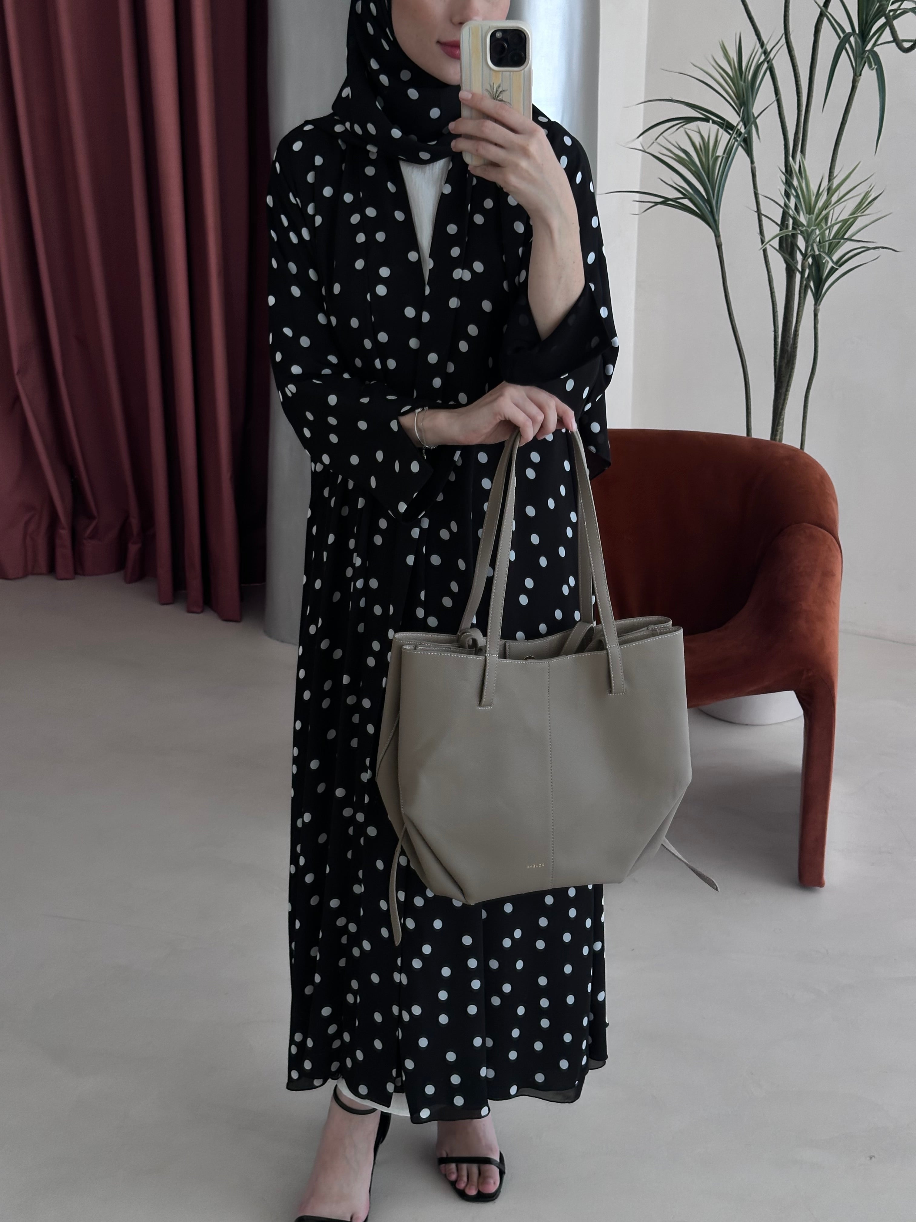 Person wearing a black polka dot dress holding a beige tote bag indoors.