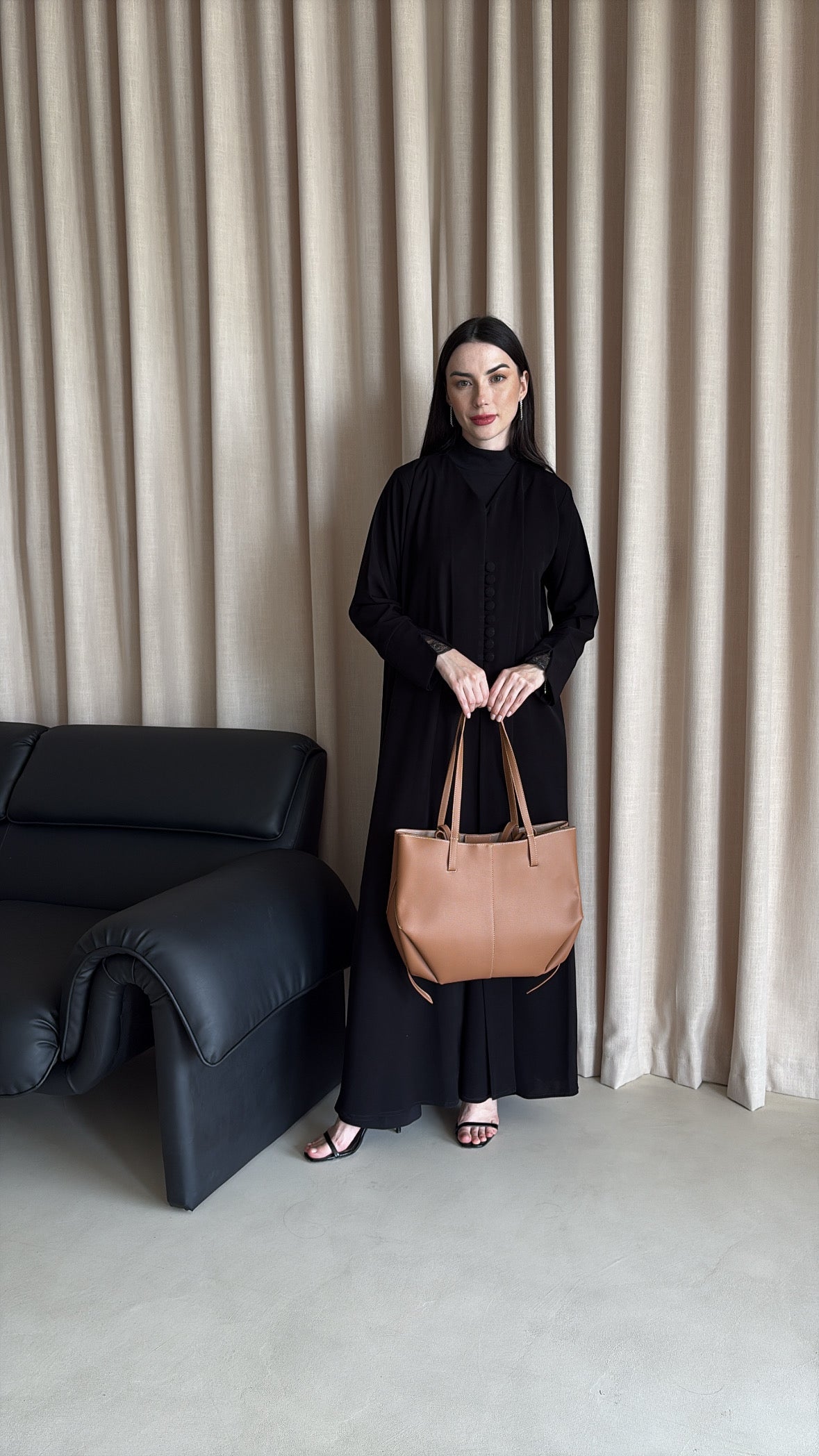 Woman holding a brown handbag in a minimalistic room with a black sofa and beige curtains.