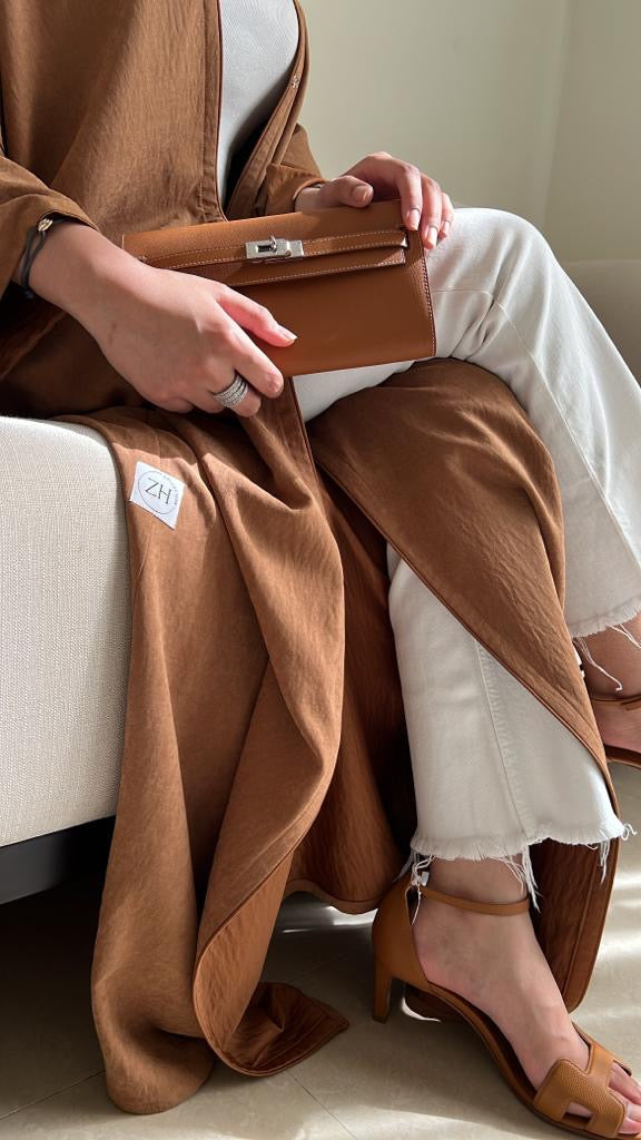 Person sitting on a couch wearing a brown coat and white pants, holding a brown handbag.