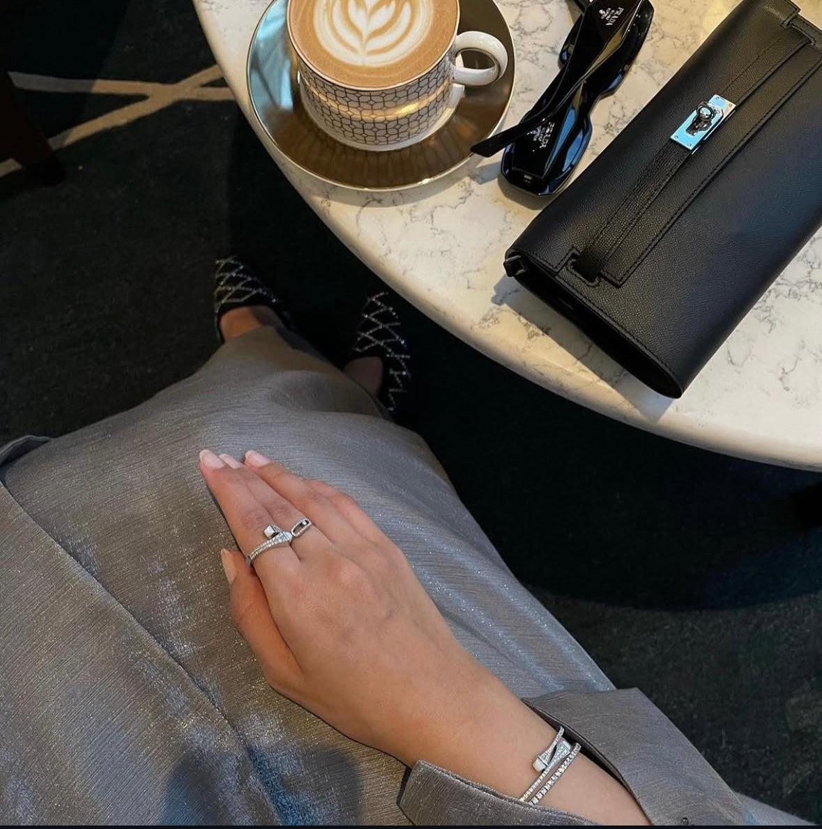 Person sitting at a table with a cup of coffee, sunglasses, and a black bag.