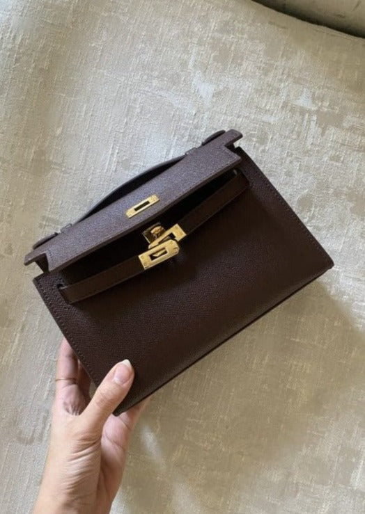 A hand holding a burgundy leather handbag with a gold clasp on a beige background.