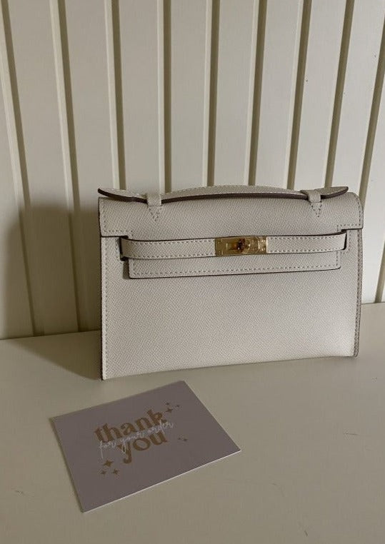A white handbag with a long strap placed on a table, with a 'thank you' card next to it.
