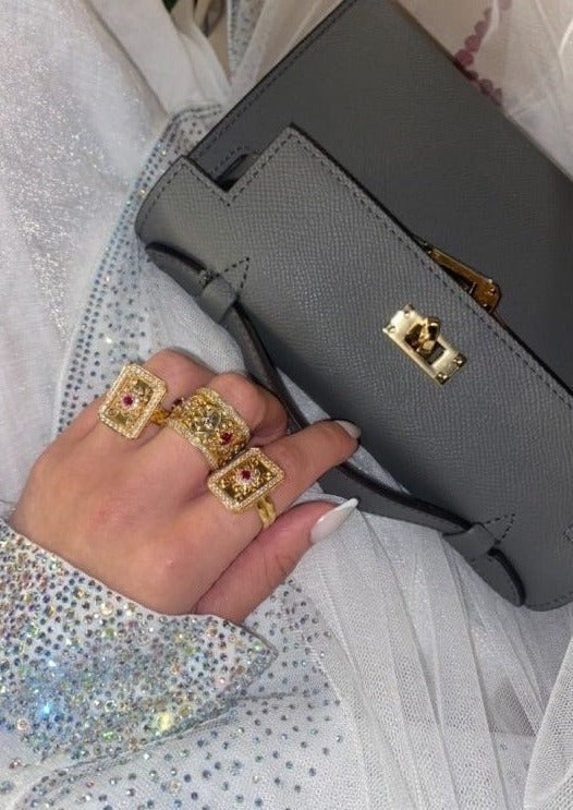 A person's hand with multiple ring accessories on their fingers, holding a grey handbag with a long strap.