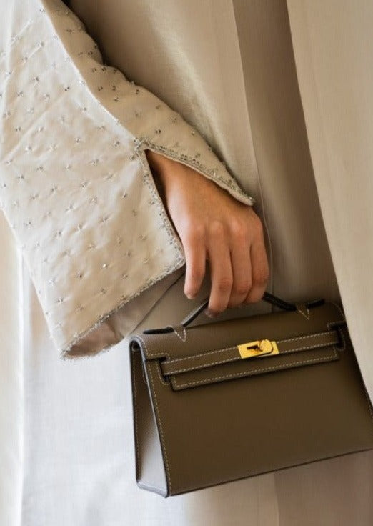 A person holding a khaki-colored handbag with a long strap and a gold-tone clasp.