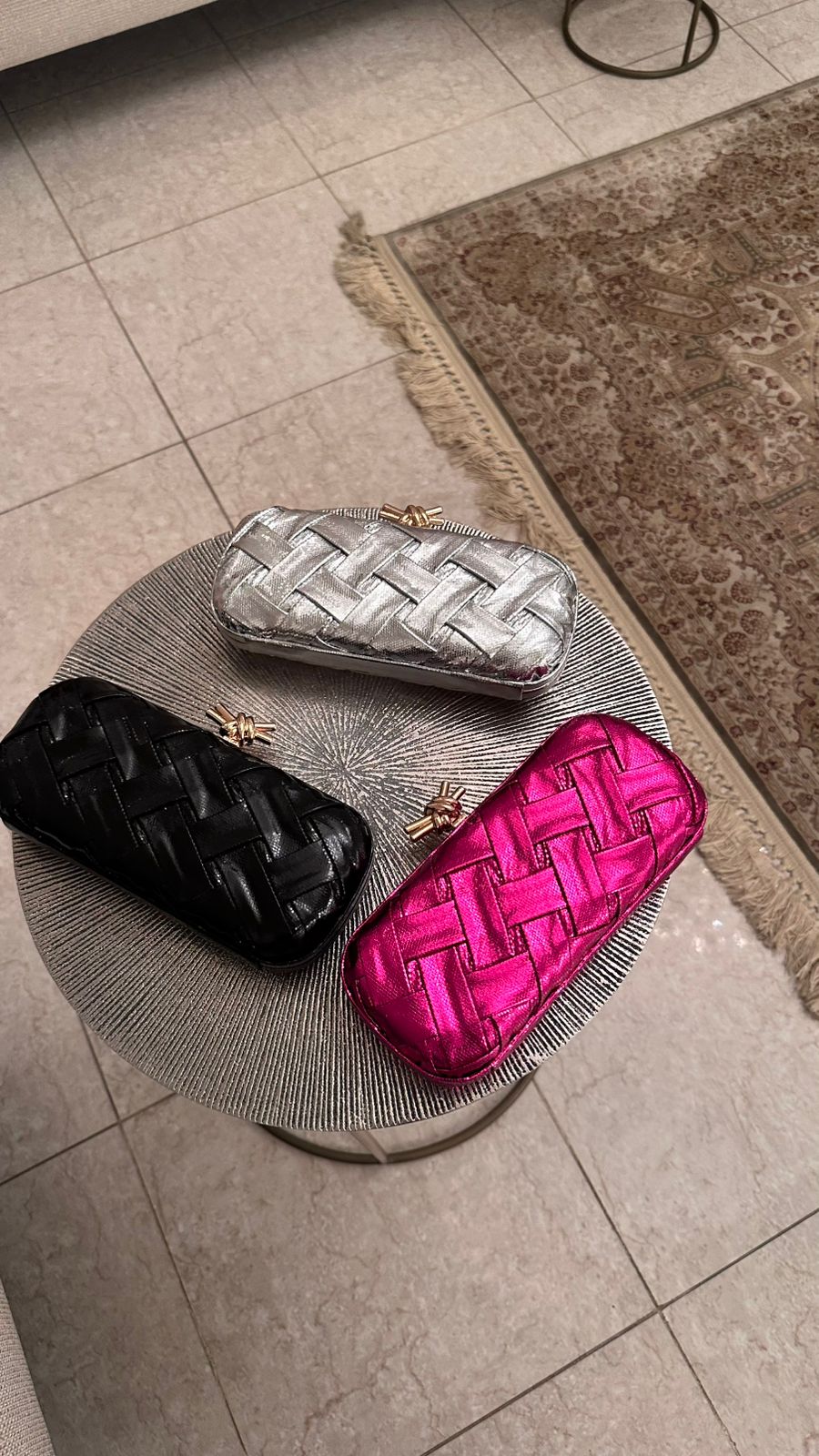 Three woven clutch bags in black, silver, and pink on a textured surface.