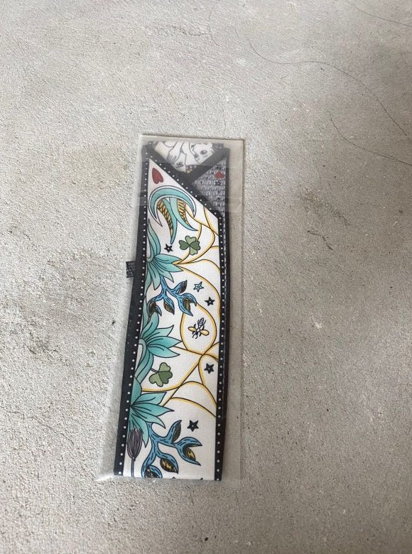 A scarf with a floral and geometric pattern, predominantly white with gold and green accents, laid out on a gray surface.