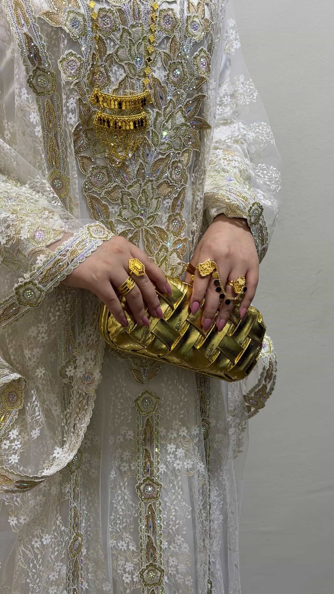 Person wearing a white embroidered outfit with gold jewelry and holding a gold clutch.