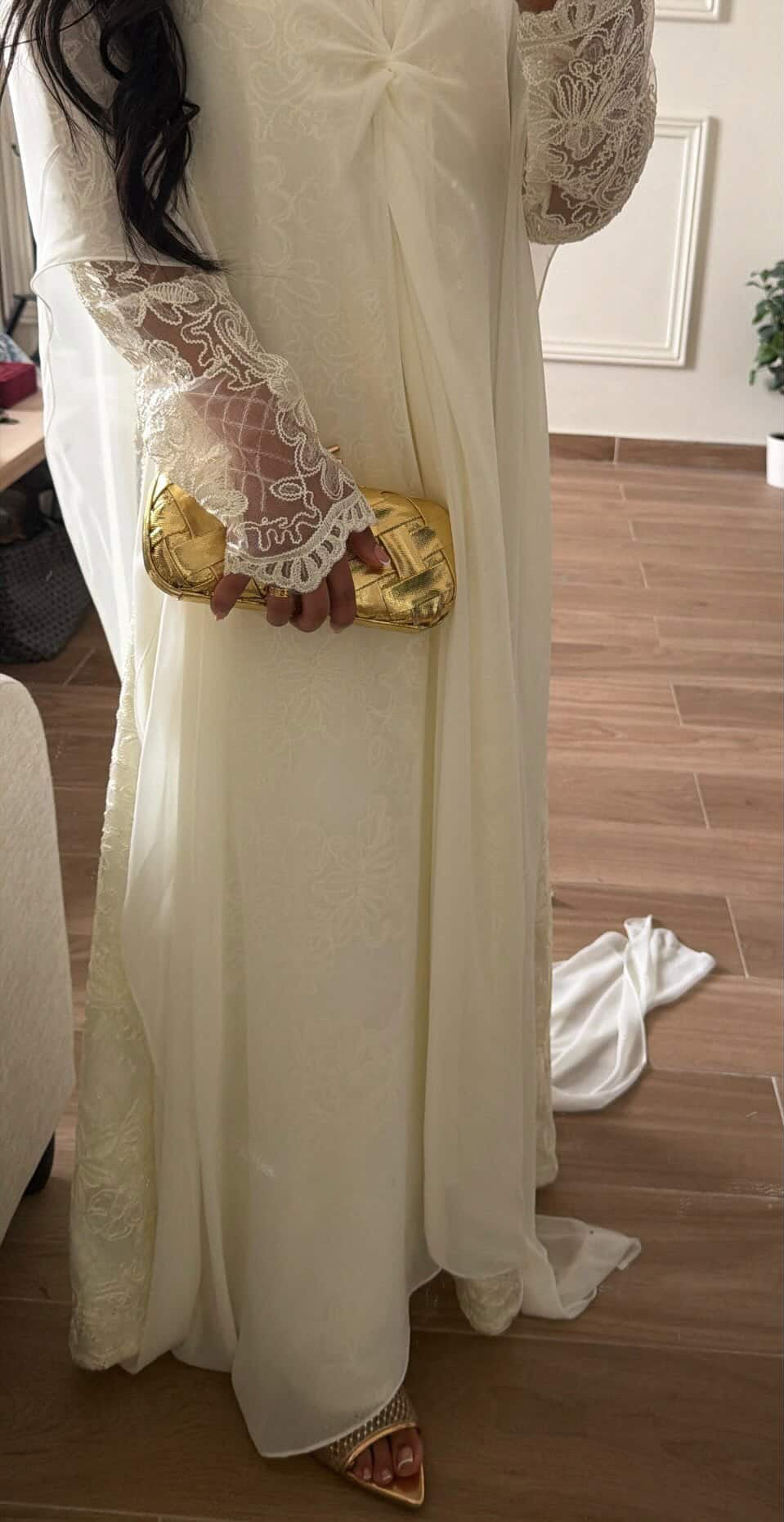 Person wearing a long white dress with lace sleeves holding a gold clutch.