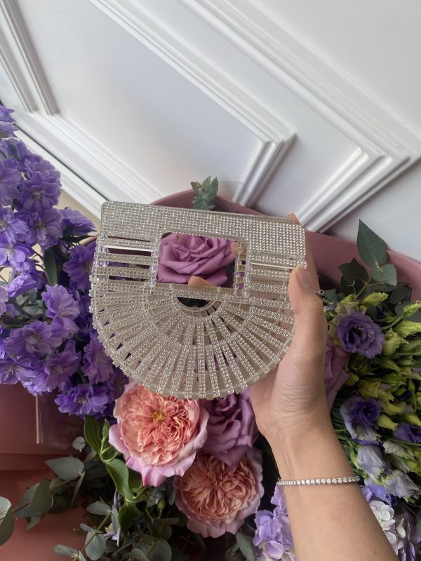A hand holding a small clear wedding crystal bag with decorative flowers in the background.