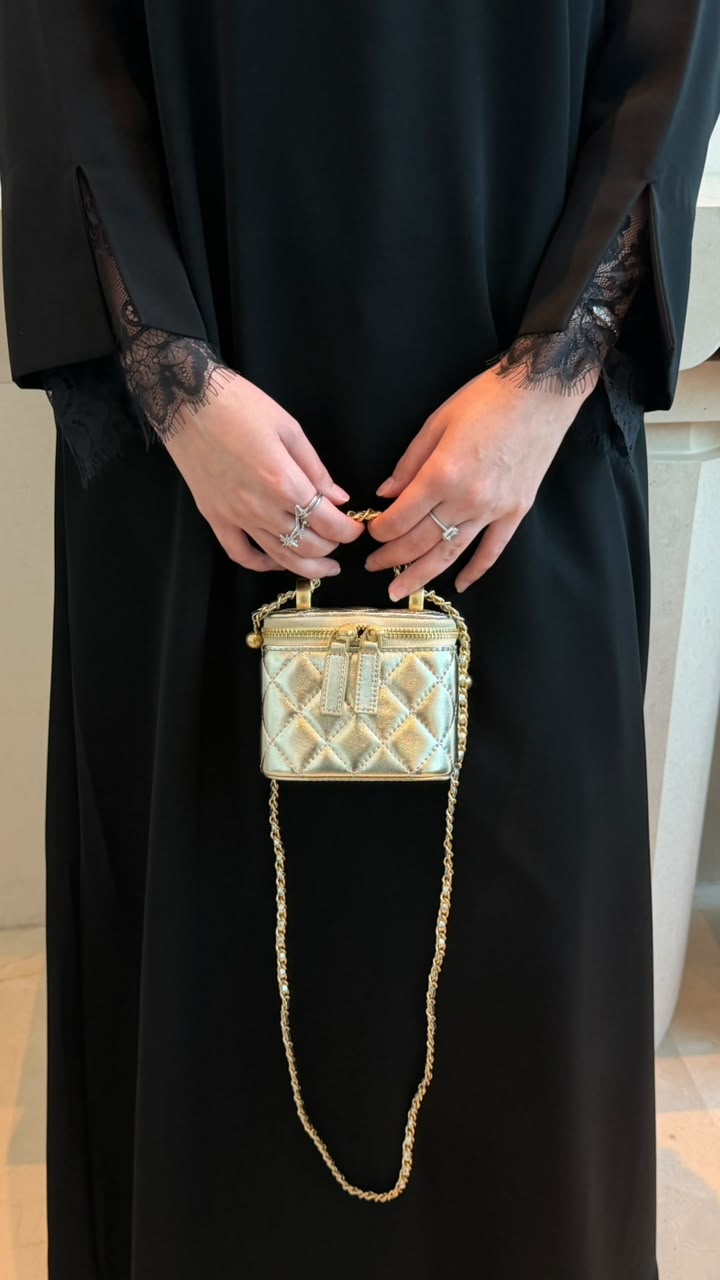 A person wearing a black abaya holds a small quilted metallic gold handbag with a gold chain strap. The person is wearing rings on both hands.