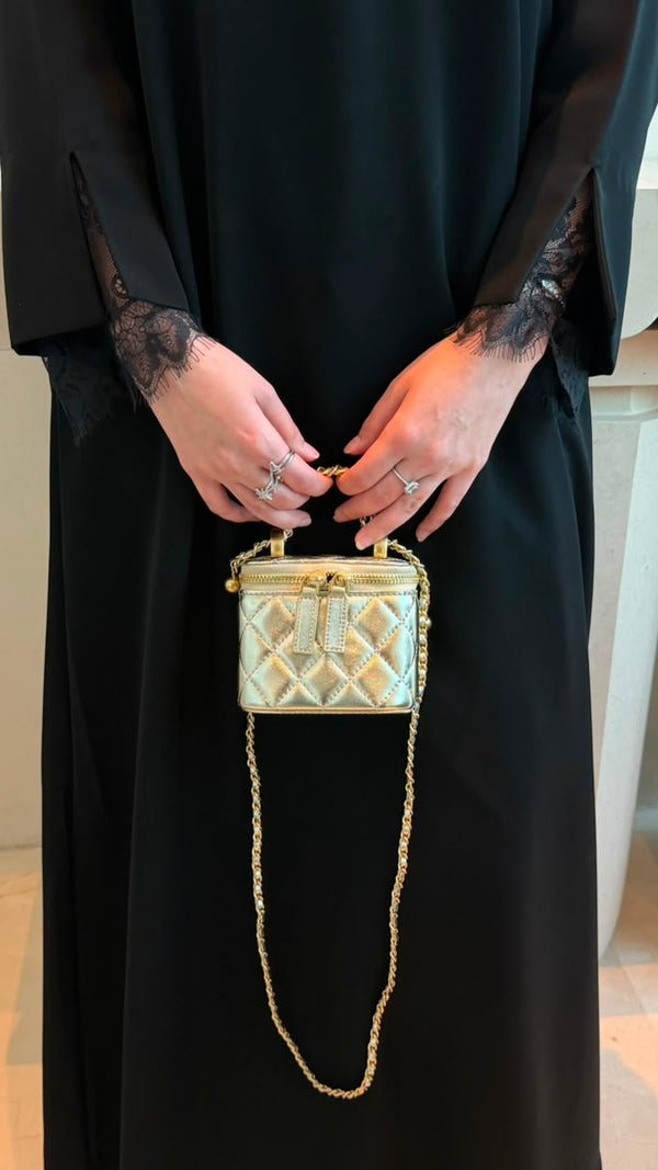 A person wearing a black abaya holds a small quilted metallic gold handbag with a gold chain strap. The person is wearing rings on both hands.
