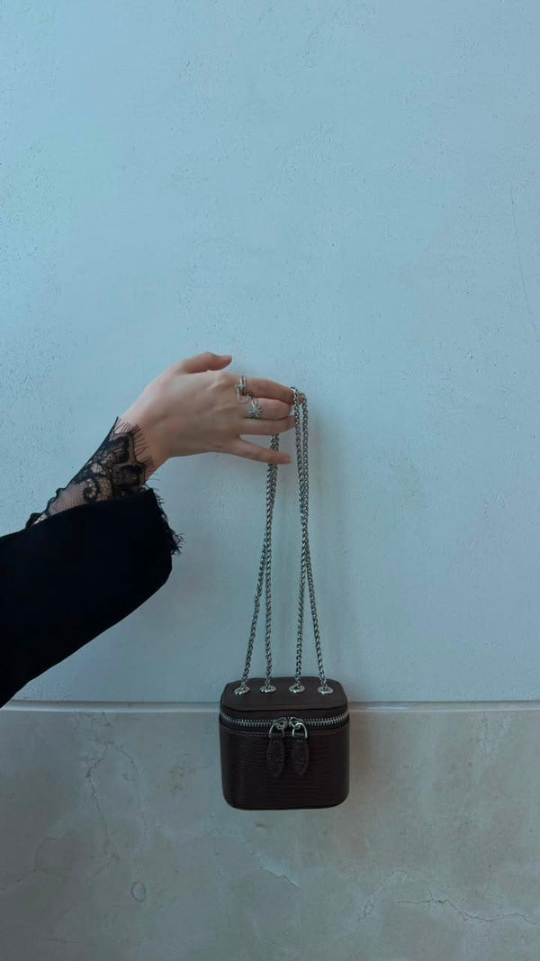 Mini Vanity Chain Bag Espresso held by chain strap by hand with black lace sleeve against light blue wall front view