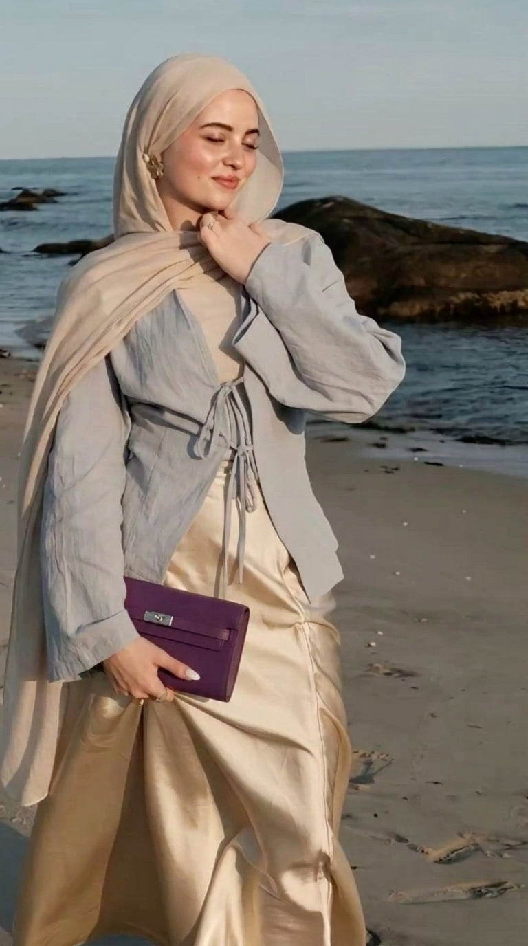 A woman wearing a beige hijab, a light blue long-sleeve tunic with a drawstring waist, and a champagne gold satin skirt walks on a sandy beach while holding a purple clutch bag.