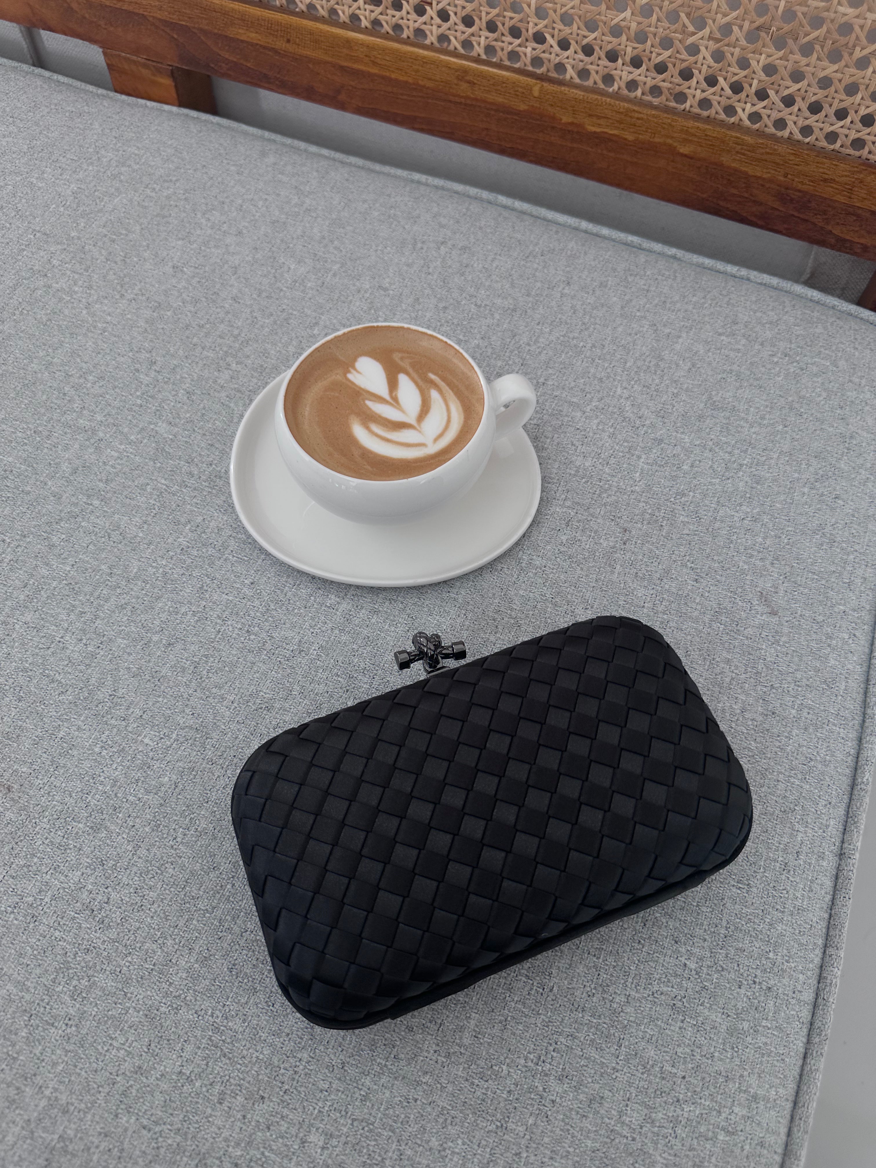 A black clutch purse with a quilted design placed on a grey surface next to a cup of coffee.