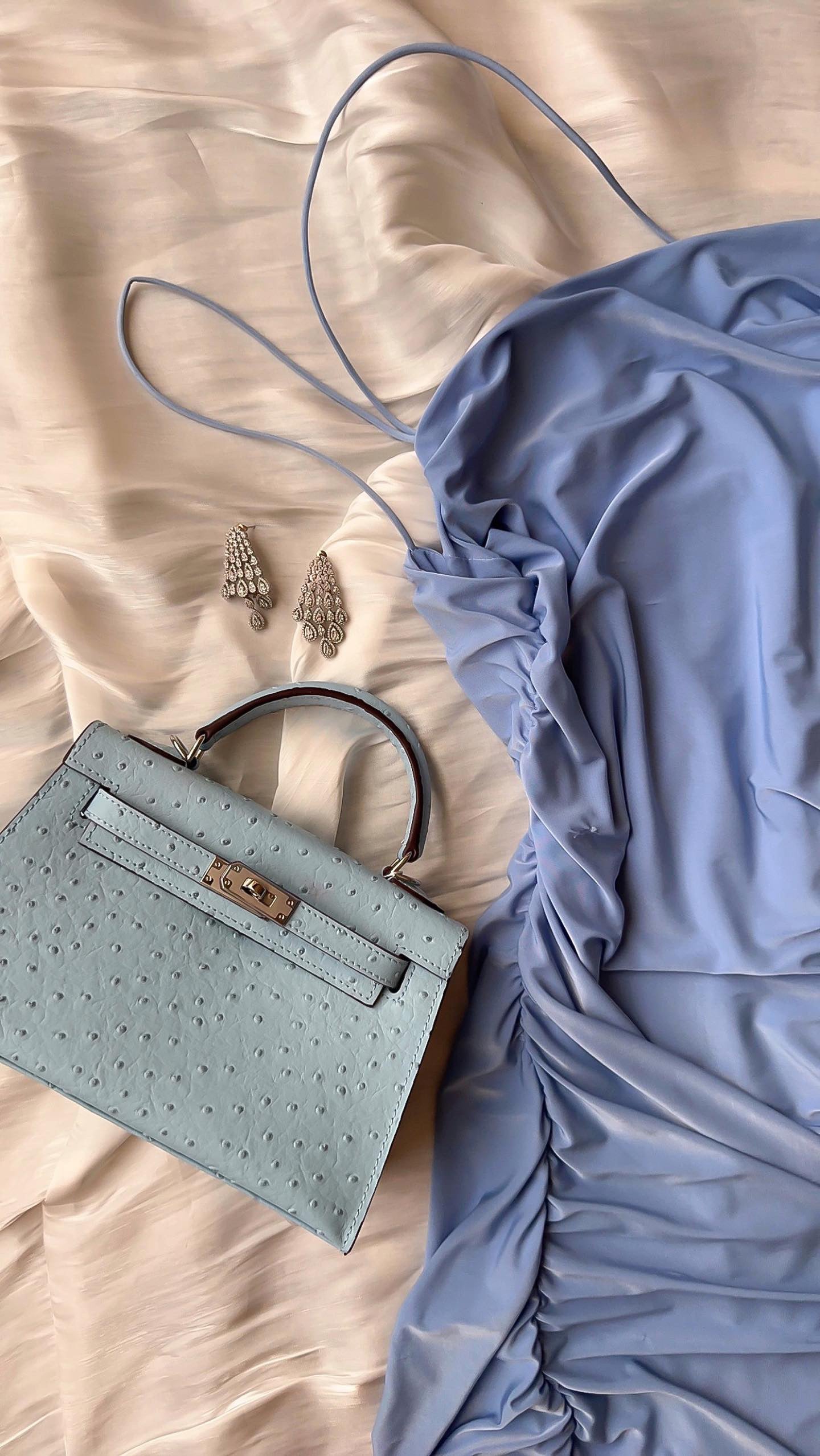 A baby blue handbag with a textured surface, alongside a long strap and some small accessories like earrings, displayed on a bed.