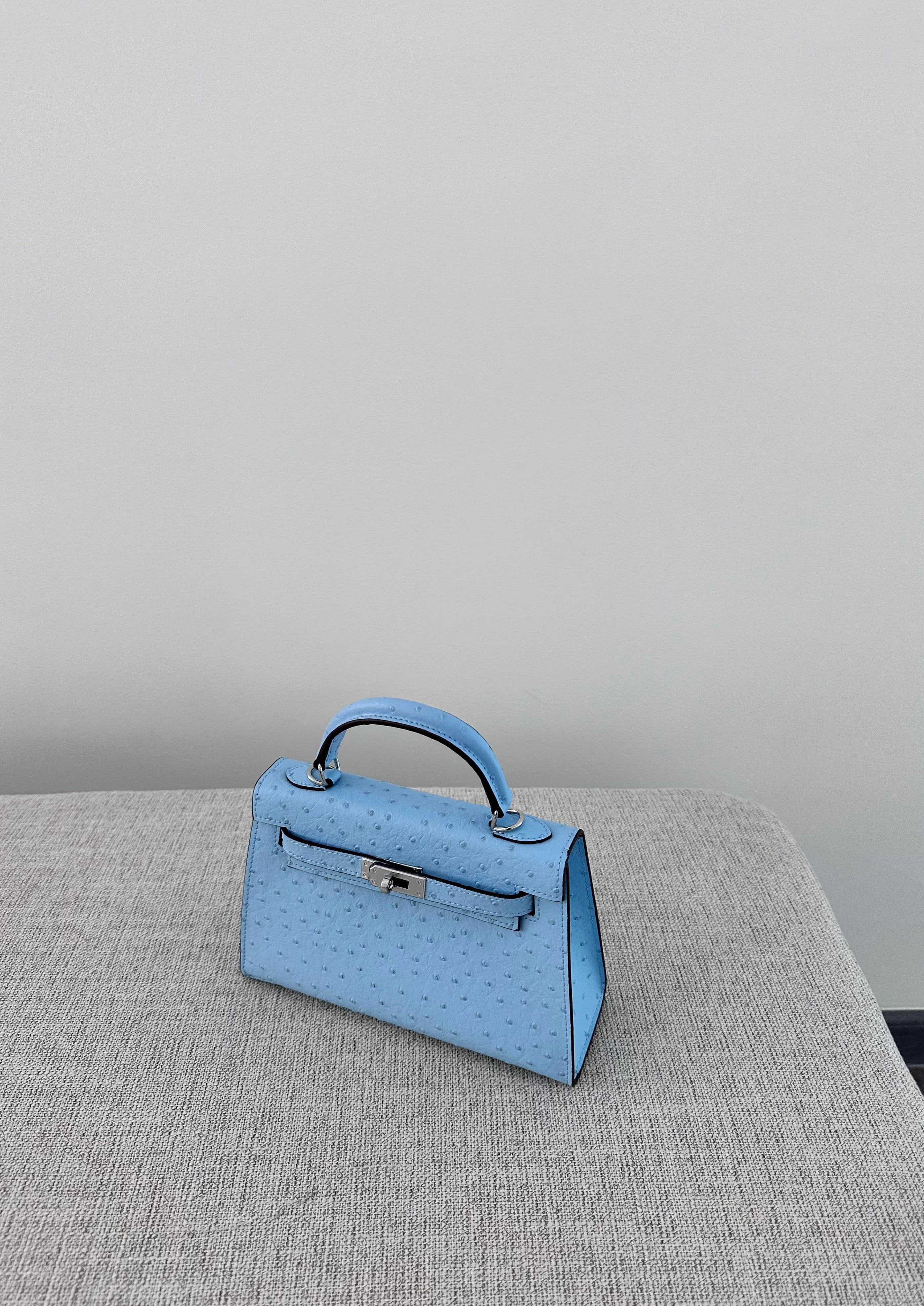 A small baby blue handbag with a clear logo and a long strap, displayed on a grey surface.
