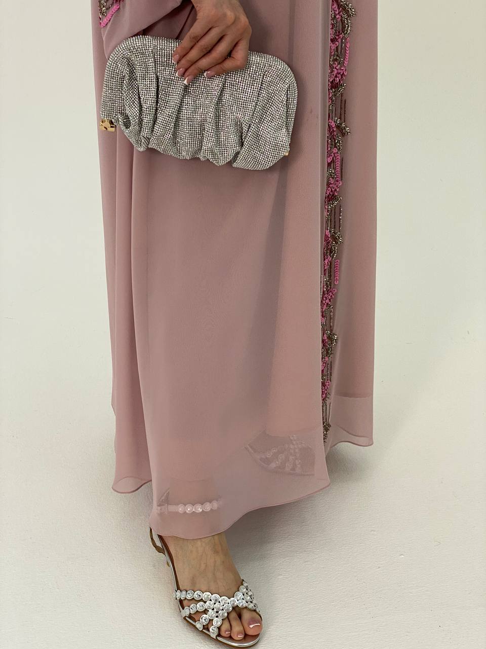 Person wearing a long light pink dress with floral details, holding a patterned clutch, and wearing pearl sandals on a white background.