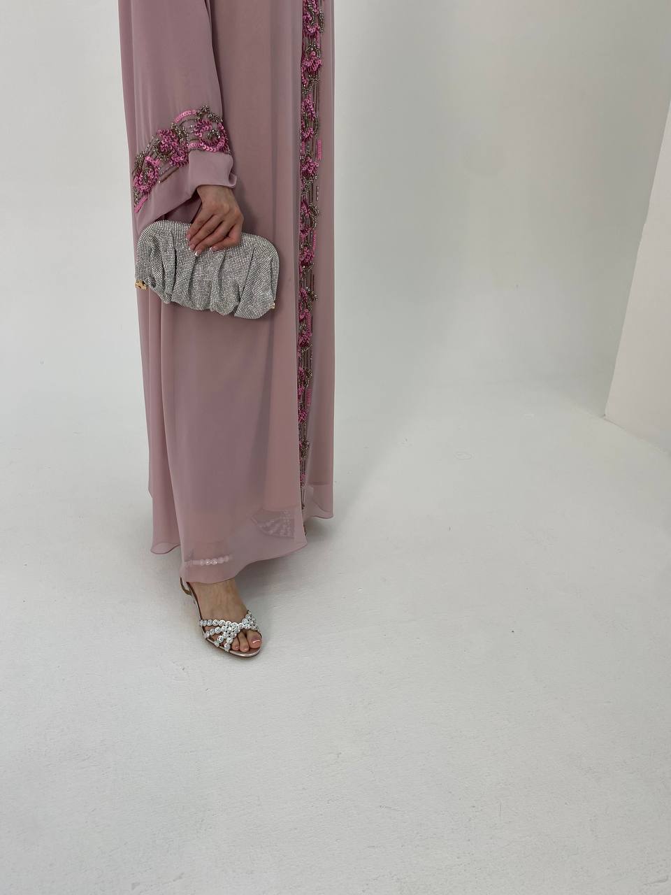 Person wearing a long, light pink dress with floral embroidery holding a gray clutch against a white background